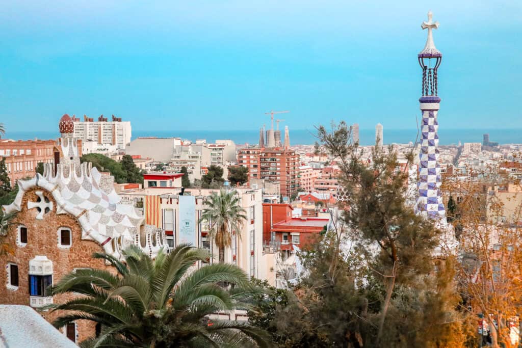 A panoramic view of Barcelona with the iconic Park Güell in the foreground featuring mosaic-tiled structures and palm trees, with the cityscape and Mediterranean Sea in the background