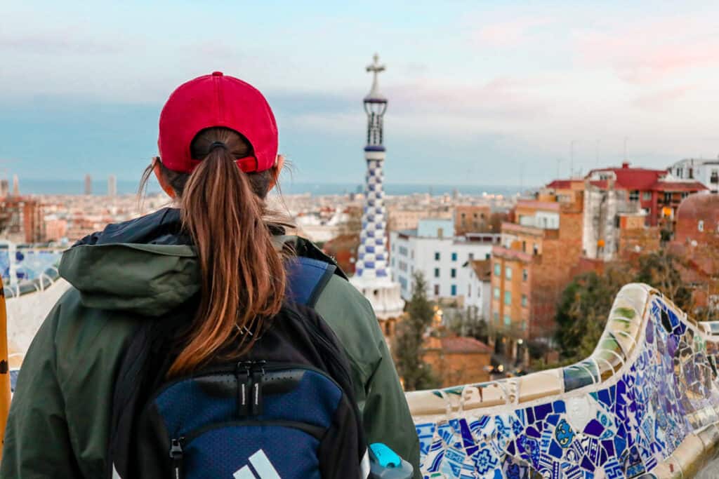 A visitor from behind wearing a red cap looks out over the colorful and intricate mosaic designs of Park Güell's balcony with a panoramic view of Barcelona.