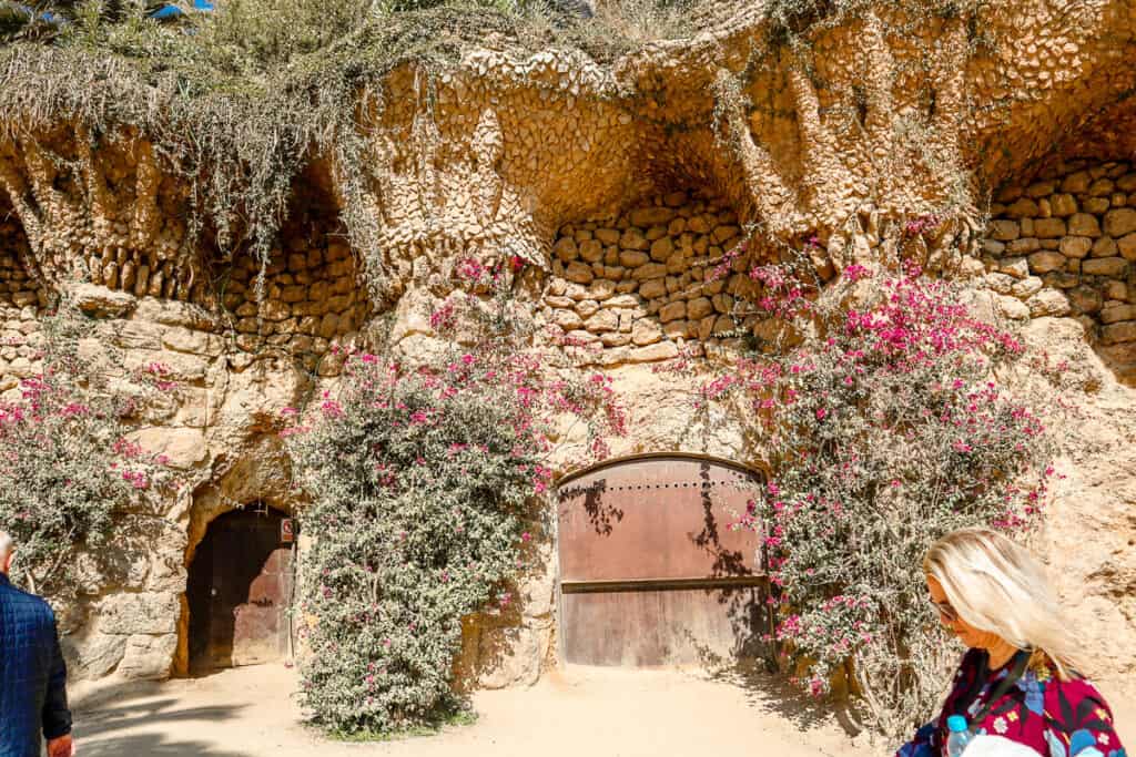A rustic stone doorway surrounded by lush pink flowers and greenery, part of Park Güell's naturalistic and organic architecture in Barcelona