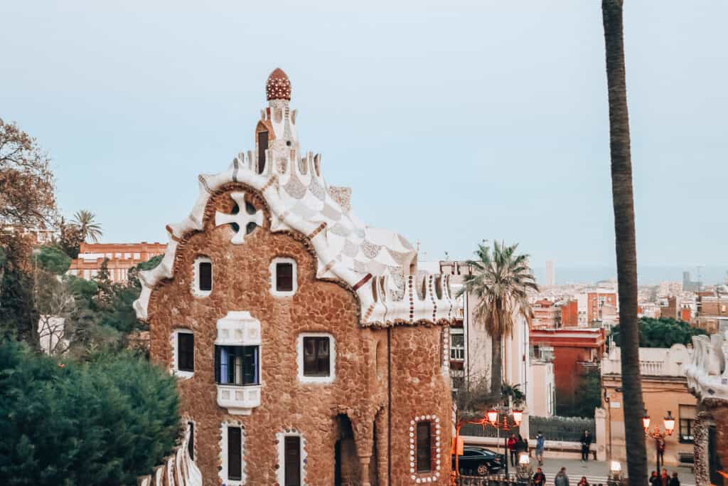 The gingerbread house-like pavilion at Park Güell in Barcelona, exhibiting Gaudí's distinctive architectural style with a whimsical mosaic roof, against a backdrop of the city and a tall palm tree