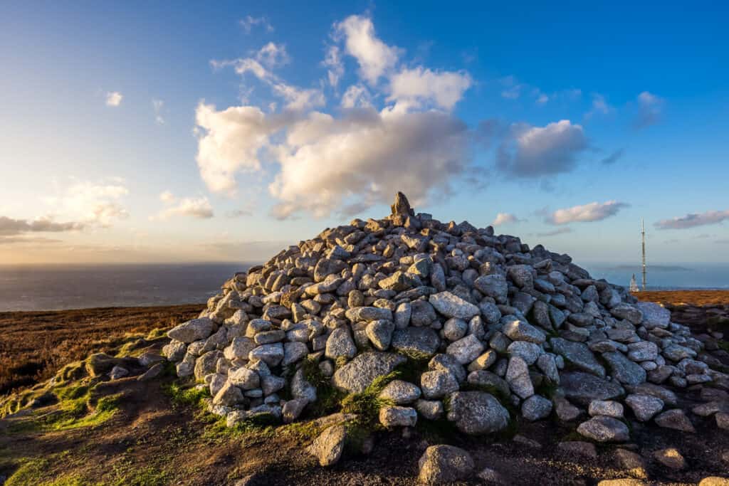 A cairn of stones at the summit of a hill under a sky with dramatic clouds, with a radio mast in the distance
