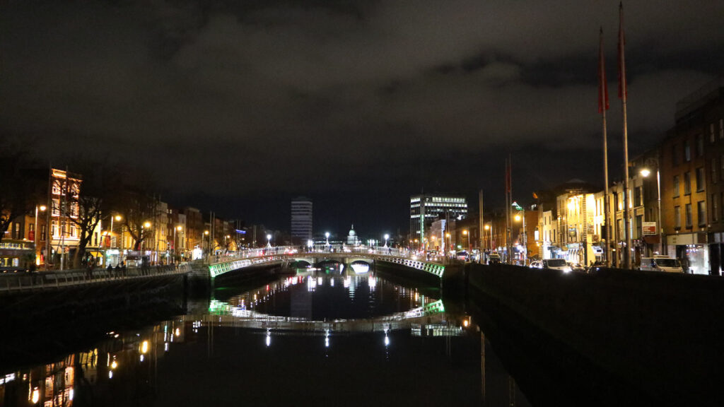 Night view of the illuminated Ha'penny Bridge over the River Liffey in Dublin, with city lights reflecting on the water.