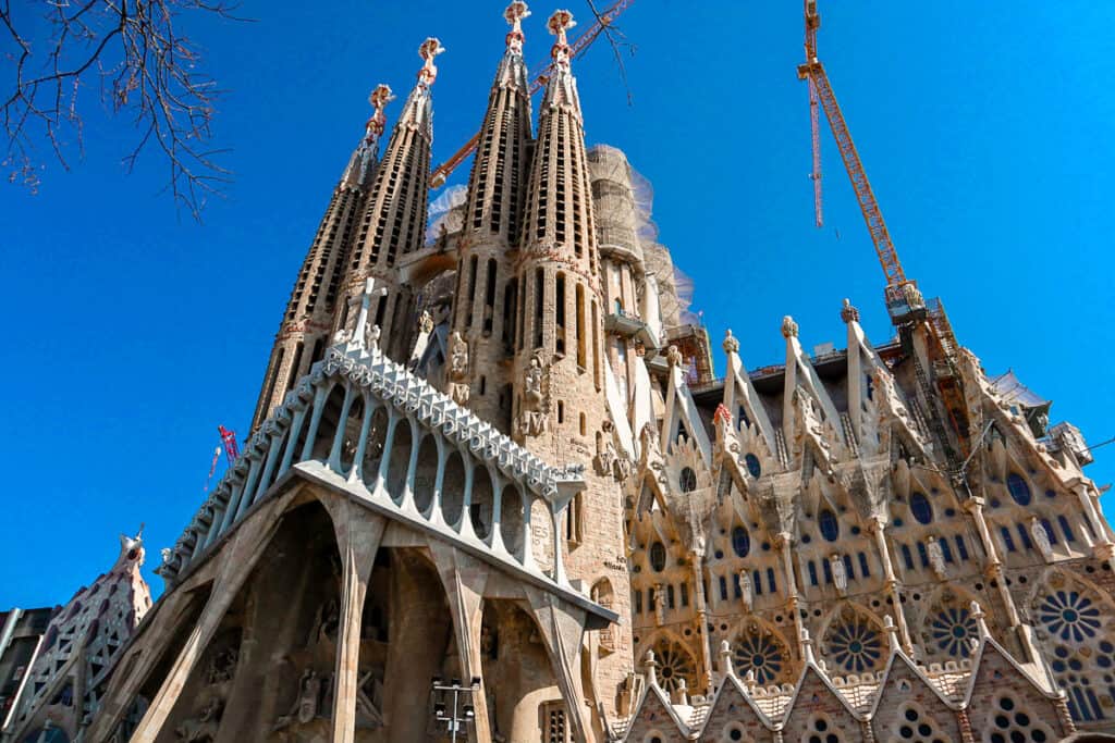 The Sagrada Familia in Barcelona, displaying its intricate spires and detailed facades, with construction cranes towering above, against a clear blue sky.