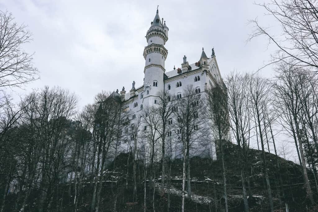 Neuschwanstein Castle looms atop a rugged cliff, its white limestone facades and tall towers contrasting with the bare trees and snowy ground in the Bavarian landscape.