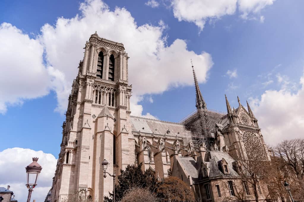 The Notre-Dame Cathedral in Paris is shown with its intricate Gothic architecture, flying buttresses, and spire, under a sky with soft clouds.