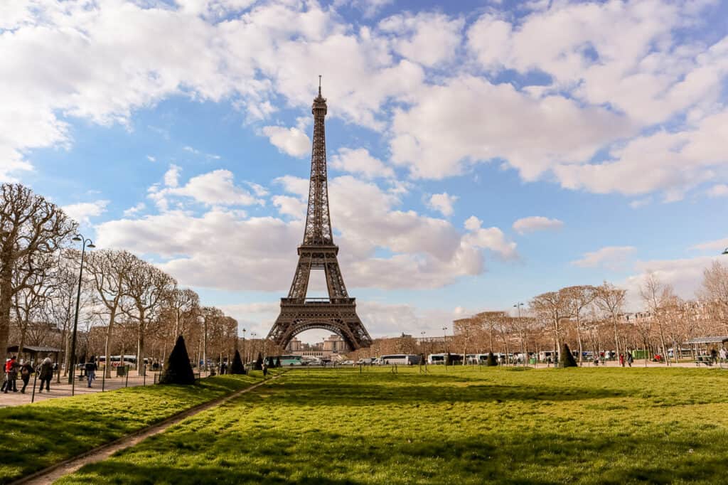 The Eiffel Tower stands tall under a blue sky dotted with clouds, flanked by leafless trees and a lush green lawn in the foreground, a scene capturing the essence of Paris.