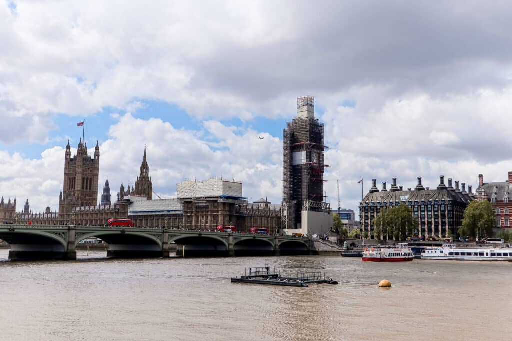 Big Ben and the Houses of Parliament in London, partially obscured by scaffolding, with the River Thames and Westminster Bridge in the foreground.