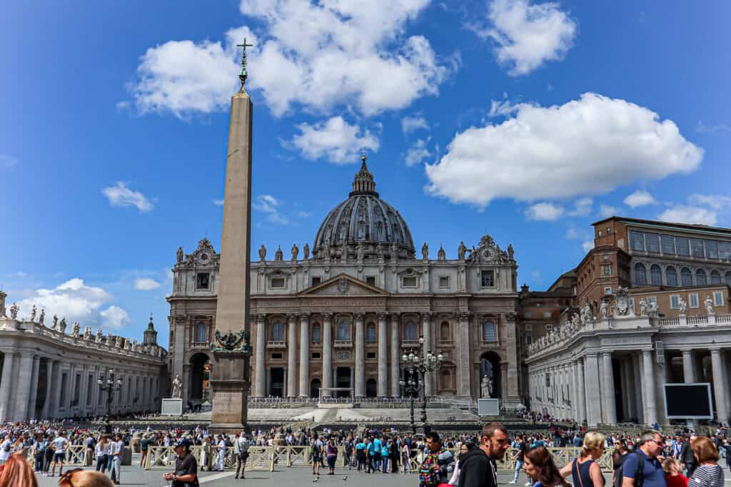 St. Peter's Square in Vatican City, crowded with visitors, showcasing the grandeur of St. Peter's Basilica with its Renaissance architecture and the iconic obelisk in the foreground.