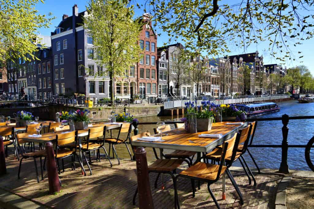 Outdoor riverside café in Amsterdam with wooden tables set up for dining, overlooking the beautiful canal and passing boats